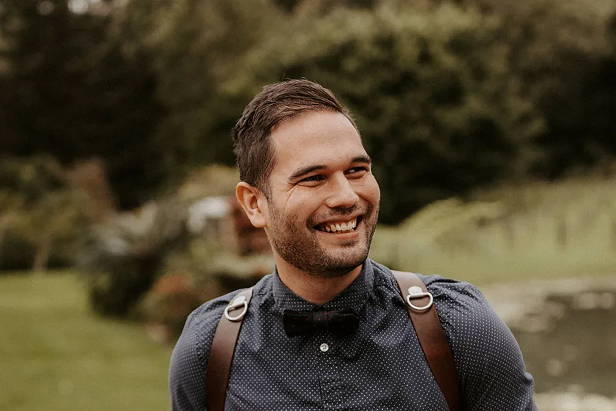 Sean Harman smiling, wearing a bow tie with two cameras on a leather harness in a garden