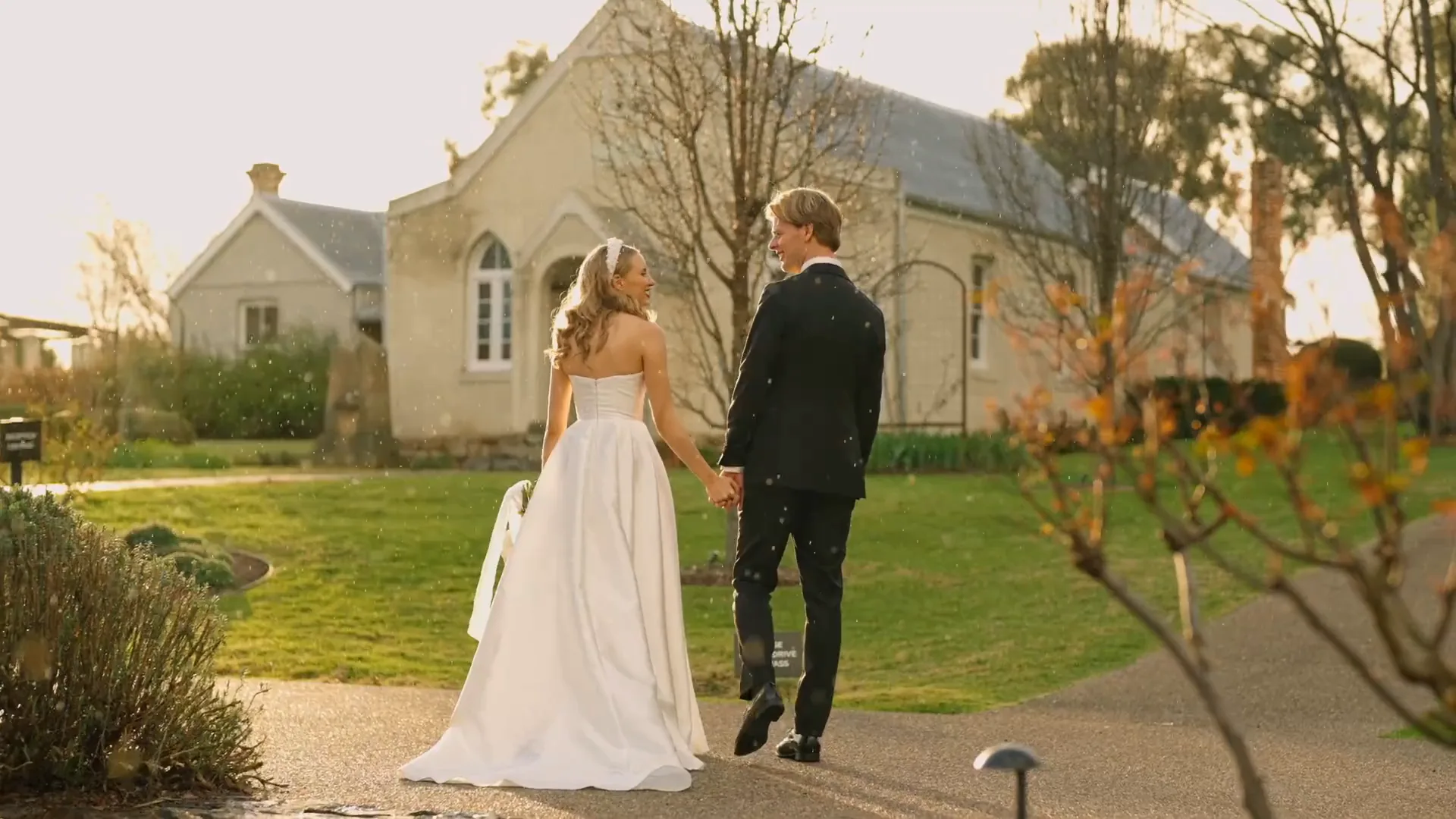 A bride and groom walking hand in hand across the lawn at golden hour